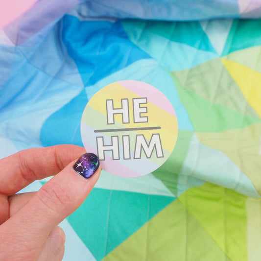 A hand holding a round pastel-colored vinyl sticker with the text 'HE HIM' written in the center, against a colorful geometric background.