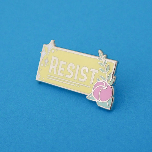 A yellow enamel pin with the word 'RESIST' written on it, featuring a flower on the right side.