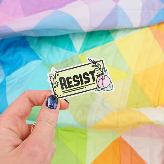 A hand holding a vinyl sticker with the word 'RESIST' on it, in front of a colorful geometric pattern.
