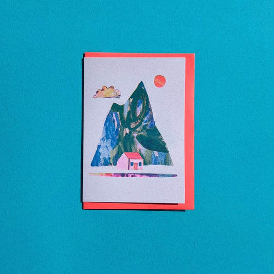 A greeting card with a colorful mountain and house design, accompanied by a red envelope, displayed against a blue background.
