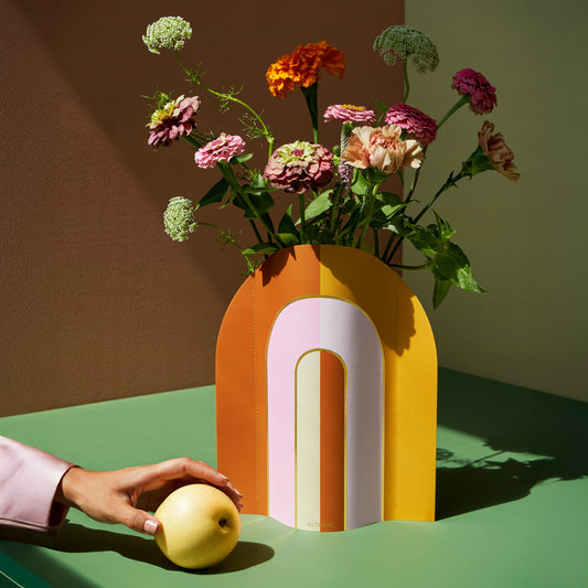 Colorful paper vase with flowers on a green surface, hand holding an apple.