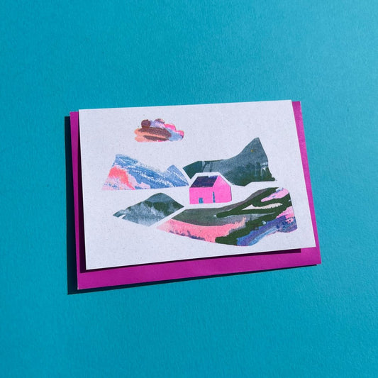 A greeting card with an illustration of a pink house with surrounding landscape 