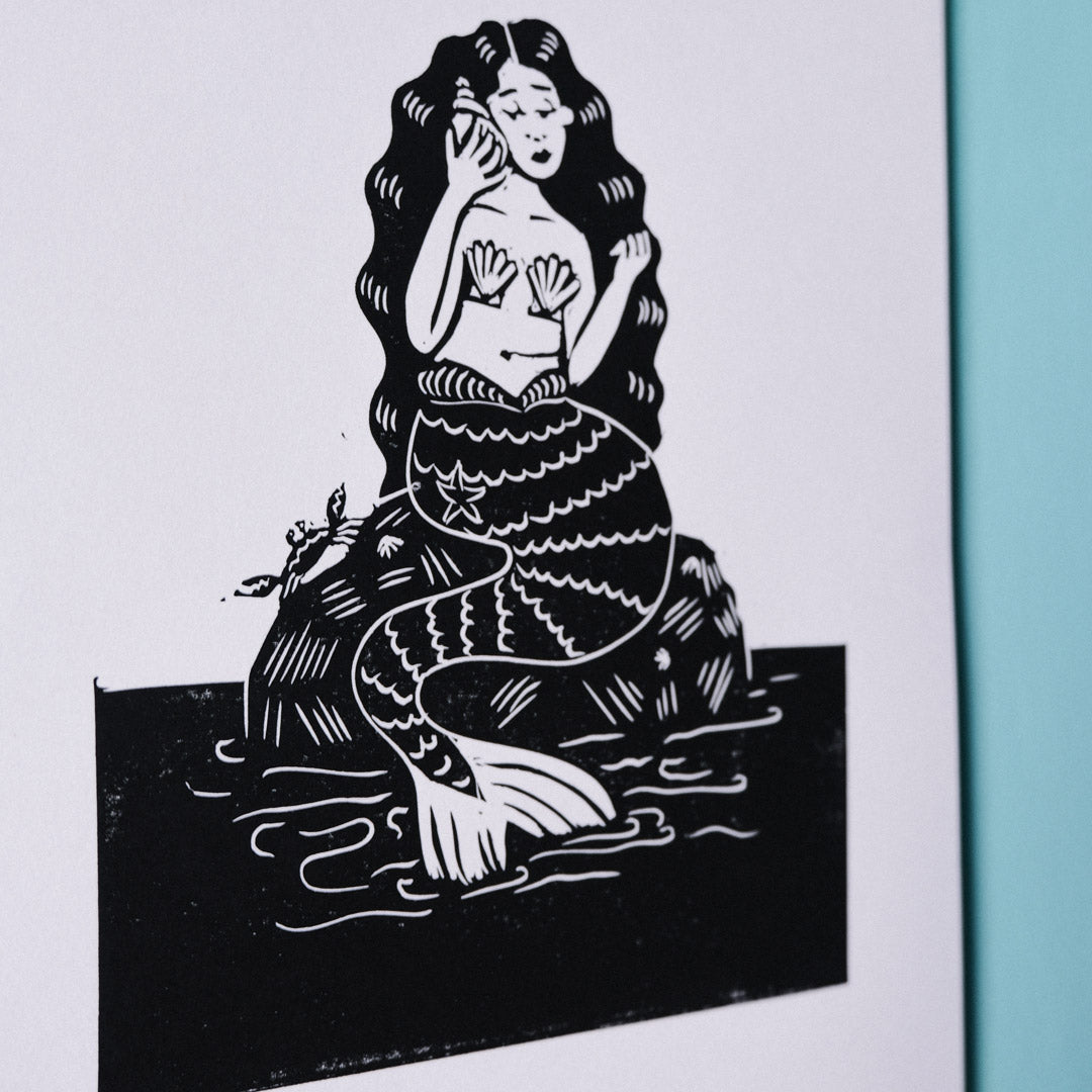 A linocut illustration of a mermaid sitting on a rock, holding a mobile phone to her ear.