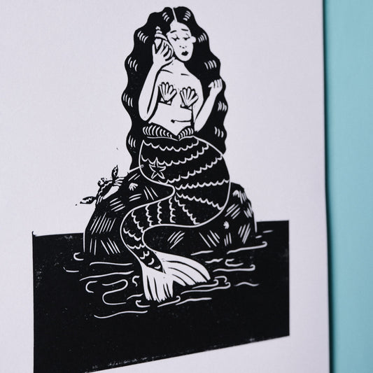 A linocut illustration of a mermaid sitting on a rock, holding a mobile phone to her ear.
